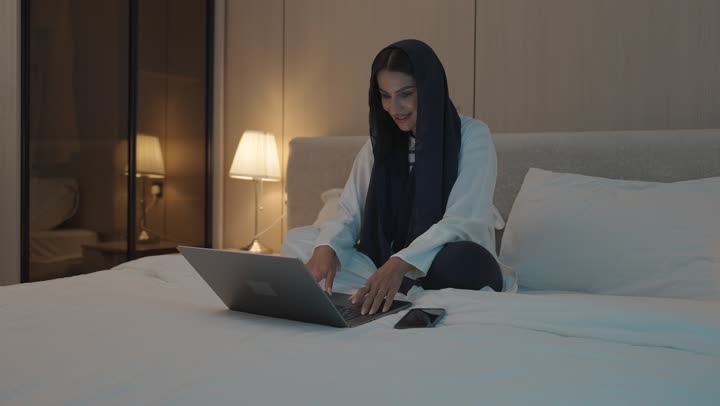 Making a video call, gestures indicating greeting and peace, a young Saudi Arabian Gulf woman sitting in the bedroom on the bed using the laptop, chatting and socializing, surfing the internet and using modern technologies, conducting a live broadcast