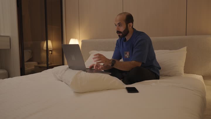 تصفح الانترنت واستخدام تقنيات حديثة, اجراء بث مباشر، القيام بمكالمة فيديو, ايماءات تدل على التحية والسلام, شاب عربي خلجي سعودي يجلس في غرفة النوم على السرير  يقوم باستخدام الحاسوب المحمول, الدردشة و التواصل الاجتماعي