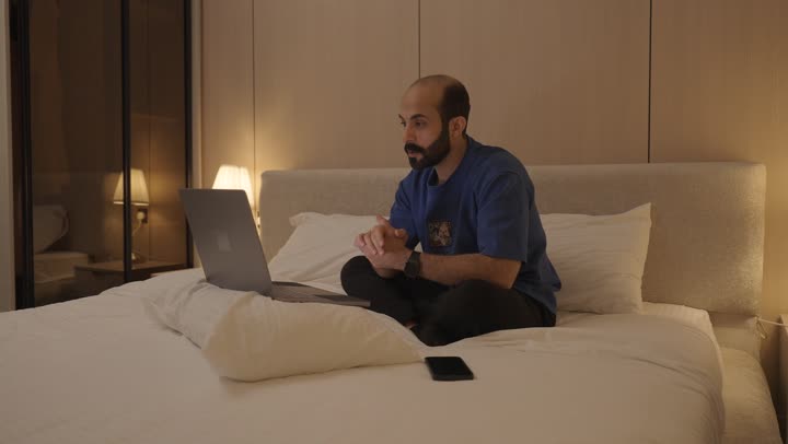 تصفح الانترنت واستخدام تقنيات حديثة, اجراء بث مباشر، القيام بمكالمة فيديو, ايماءات تدل على التحية والسلام, شاب عربي خلجي سعودي يجلس في غرفة النوم على السرير  يقوم باستخدام الحاسوب المحمول, الدردشة و التواصل الاجتماعي