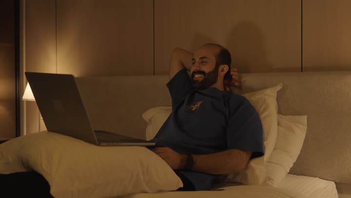 Browsing the internet and using modern technologies, conducting a live broadcast, making a video call, gestures indicating greetings and peace, a young Saudi Arabian Gulf man sitting in the bedroom on the bed using the laptop, chatting and socializing
