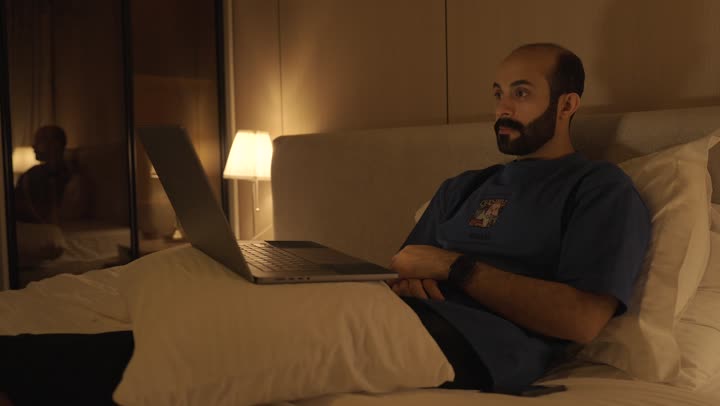 Browsing the internet and using modern technologies, conducting a live broadcast, making a video call, gestures indicating greetings and peace, a young Saudi Arabian Gulf man sitting in the bedroom on the bed using the laptop, chatting and socializing