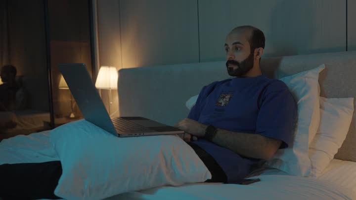 Browsing the internet and using modern technologies, conducting a live broadcast, making a video call, gestures indicating greetings and peace, a young Saudi Arabian Gulf man sitting in the bedroom on the bed using the laptop, chatting and socializing