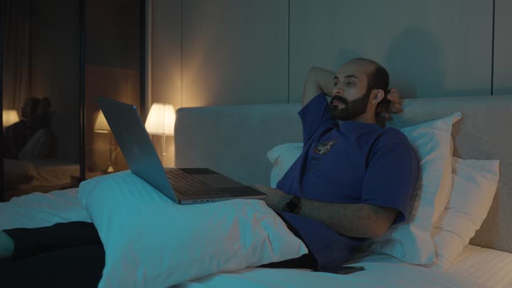 تصفح الانترنت واستخدام تقنيات حديثة, اجراء بث مباشر، القيام بمكالمة فيديو, ايماءات تدل على التحية والسلام, شاب عربي خلجي سعودي يجلس في غرفة النوم على السرير يقوم باستخدام الحاسوب المحمول, الدردشة و التواصل الاجتماعي