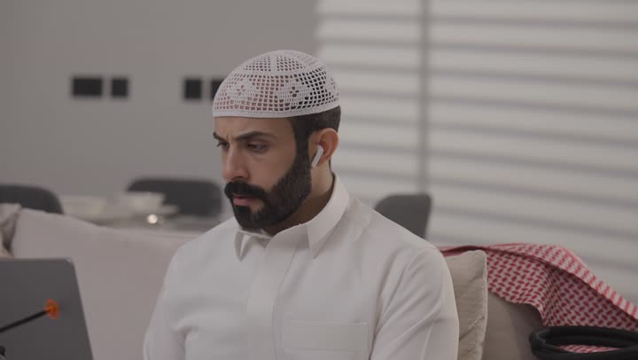 العمل عن بعد في المنزل، عائلة عربية خليجية سعودية تقضي اوقات ممتعة في غرفة المعيشة، رجل سعودي يرتدي الثوب التقليدي يقوم بانجاز المهام عبر الحاسوب المحمول بايماءات التركيز