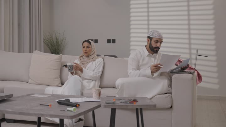 العمل عن بعد في المنزل، عائلة عربية خليجية سعودية تقضي اوقات ممتعة في غرفة المعيشة، رجل سعودي يرتدي الثوب التقليدي يقوم بانجاز المهام عبر الحاسوب المحمول بايماءات التركيز
