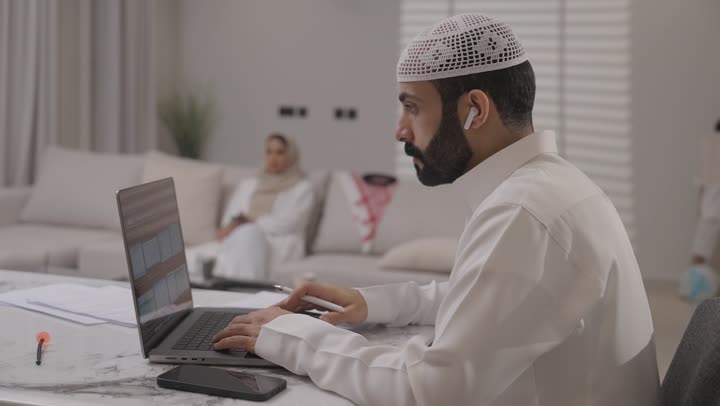 العمل عن بعد في المنزل، عائلة عربية خليجية سعودية تقضي اوقات ممتعة في غرفة المعيشة، رجل سعودي يرتدي الثوب التقليدي يقوم بانجاز المهام عبر الحاسوب المحمول بايماءات التركيز