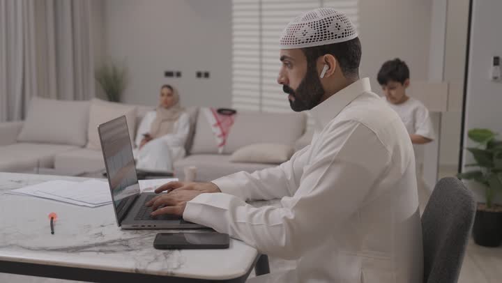 العمل عن بعد في المنزل، عائلة عربية خليجية سعودية تقضي اوقات ممتعة في غرفة المعيشة، رجل سعودي يرتدي الثوب التقليدي يقوم بانجاز المهام عبر الحاسوب المحمول بايماءات التركيز