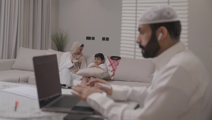 Working remotely from home, a Saudi Arabian Gulf family having fun in the living room, a Saudi man wearing traditional thobe completing tasks on his laptop with focus gestures