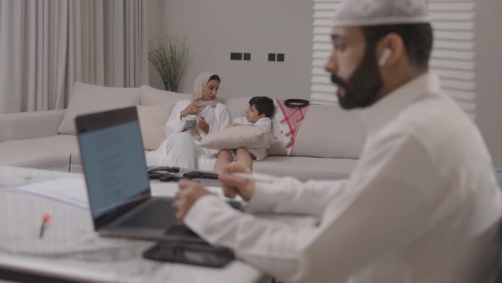 العمل عن بعد في المنزل، عائلة عربية خليجية سعودية تقضي اوقات ممتعة في غرفة المعيشة، رجل سعودي يرتدي الثوب التقليدي يقوم بانجاز المهام عبر الحاسوب المحمول بايماءات التركيز