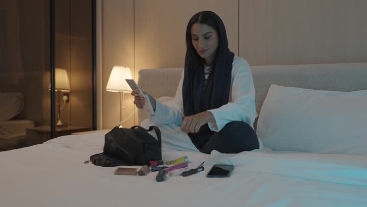 أدوات تجميل نسائيه عصريه، إمرأة عربية خليجية سعودية تجلس على السرير في غرفة النوم تقوم بوضع الميكاب، مفهوم الانوثه والاهتمام بالمظهر الخارجي, الاستعداد والتجهز للخروج
