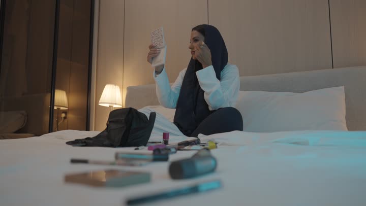 أدوات تجميل نسائيه عصريه، إمرأة عربية خليجية سعودية تجلس على السرير في غرفة النوم تقوم بوضع الميكاب، مفهوم الانوثه والاهتمام بالمظهر الخارجي, الاستعداد والتجهز للخروج