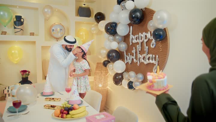 Feelings of happiness, love and tenderness in a small family gathering, sudden and amazed gestures, happy holidays and occasions, sharing a wonderful family atmosphere inside the home, an Arab Gulf mother presenting a birthday cake decorated with candles, an Arab Gulf Emirati family celebrating their daughter's birthday
