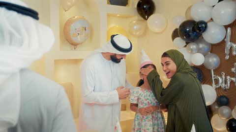 التعبير عن المحبة والتقدير، مشاعر الحب والحنان في اجتماع عائلي سعيد، مشاركة لأجواء رائعة لعائلة اماراتية داخل المنزل،عائله عربيه خليجيه اماراتية ترتدي الزي التقليدي تقوم بالاحتفال بعيد ميلاد طفلتهم بايماءات السعادة و السرور، رجل خليجي يقوم بتقديم الهديه للطفلة