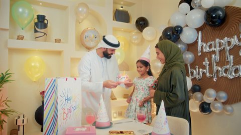 أجواء عائلية سعيدة، رجل عربي خليجي امارتي يقف بجانب زوجته ويحمل بيده قالب كيك يحتفلون بابنتهم بإيماءات تدل على السعادة والمحبة، مفهوم الأسرة والطفل، قضاء أوقات عائلية ممتعة