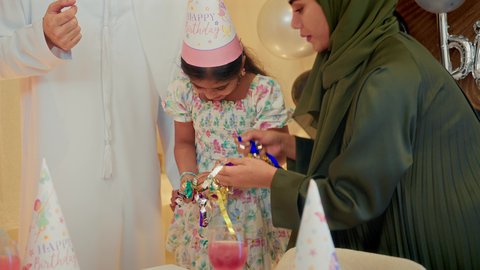 الاحتفال بأعياد الميلاد في أجواء عائلية دافئة، تعليق الزينة والبالونات الملونة، إيماءات السعادة والمفجاءة، استقبال الهدايا من الحضور والأهل،تصوير مقرب لطفلة عربية خليجية اماراتية ترتدي فستان تحتفل بعيد ميلادها برفقة أسرتها