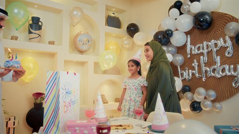 إيماءات السعادة والسرور باستقبال الهدايا من الحضور والأهل، تعليق الزينة والبالونات الملونة،  الاحتفال بأعياد الميلاد في أجواء عائلية دافئة، طفلة عربية خليجية اماراتية ترتدي فستان تحتفل بعيد ميلادها برفقة أسرتها