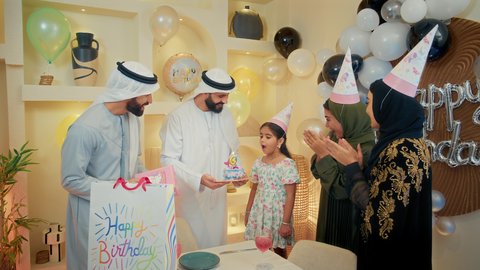 قضاء أوقات عائلية ممتعه، عائله عربيه خليجيه اماراتية ترتدي الملابس التقليدية يقومان بالاحتفال بعيد ميلاد ابنتهما الصغيرة، فتاة ترتدي فستان ملون تقوم باطفاء شمعه كيكه العيد ميلاد بايماءات السعادة و السرور