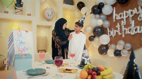 تعزيز روابط الامومه والحنان، تعليق الزينة والبالونات الملونة، الاحتفال بأعياد الميلاد ضمن أجواء عائلية، ام عربيه خليجيه اماراتية ترتدي العباءه تقوم بالاحتفال و تقبيل ابنها بايماءات السعادة و السرور