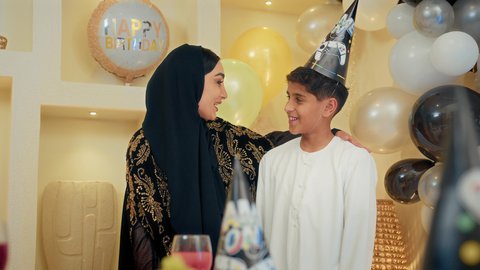 Hanging colorful decorations and balloons, celebrating birthdays in a family atmosphere, an Emirati Gulf Arab mother wearing an abaya celebrating and kissing her son with gestures of happiness and pleasure, strengthening the bonds of motherhood and tenderness