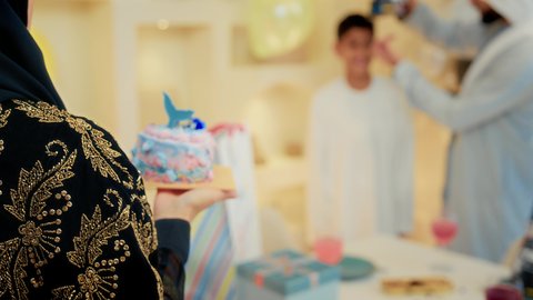 مفهوم الإهداء والتقدير، زوجان عربيان خليجيان اماراتيان يرتديان الزي التقليدي يقومان بالاحتفال بعيد ميلاد ابنهم، تصوير مقرب لام عربية خليجيه تقوم تقديم الكعكة الشهية والهدايا، إيماءات الفرح و السعادة، مشاعر الحب والحنان في اجتماع عائلي سعيد