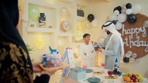وضع الزينة والبالونات الملونة واكسسوارات الحفلة، زوجان عربيان خليجيان اماراتيان يرتديان الزي التقليدي يقومان بالاحتفال بعيد ميلاد ابنهم، ام عربية خليجيه تقوم بتقبيل ابنها، إيماءات الفرح و السعادة، مشاعر الحب والحنان في اجتماع عائلي سعيد