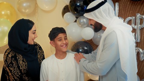 تبادل المحبة والمودة بين الآباء والأبناء، زوجان عربيان خليجيان اماراتيان يرتديان الزي التقليدي يقومان بتقبيل ابنهم، عائلة عربية خليجية إماراتية تقوم بالاحتفال بعيد الميلاد، إيماءات الفرح و السعادة، مشاعر الحب والحنان في اجتماع عائلي سعيد