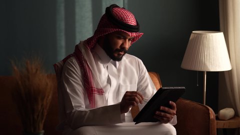 العمل عن بعد، مهنة ووظيفة مكتبية، استخدام التقنية الحديثه المتطورة في الحياة العملية، ايماءات الاندماج والتركيز، رجل عربي خليجي سعودي يرتدي الثوب التقليدي والشماغ يجلس على الكنبة ويعمل على الجهاز اللوحي في صالة المنزل