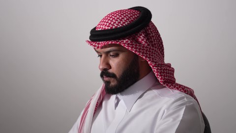مهنة ووظيفة مكتبية، العمل عن بعد، استخدام التقنية الحديثه المتطورة في الحياة العملية، ايماءات الاندماج والتركيز، بورتريه لرجل عربي خليجي سعودي يرتدي الثوب التقليدي والشماغ يقوم بالعمل على الحاسب المحمول في مقر الشركة، خلفية بيضاء