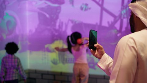رجل اماراتي يوثق اللحظات التذكاريه السعيده لابنائه عبر الجوال ،  قضاء اوقات ممتعه مع العائله ،  مراكز الالعاب الترفيهيه في الامارات،تصوير لطفلان اشقاء عربيان خليجيان إماراتيان يرتديان الزى الكاجوال ، يستمتعان بلعبة الرمي التفاعلية