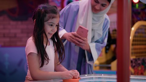 استخدام الهاتف المحمول والتكنولوجيا الحديثه ، امراه اماراتيه توثق اللحظات التذكاريه السعيده لابنتها ،طاولة مضيئة بألوان زاهية ، قضاء اوقات ممتعه مع العائله ، مراكز الالعاب الترفيهيه في الامارات، تصوير لطفلة عربية خليجية إماراتية ترتدي الزى الكاجوال ، تستمتع بلعب الهوكي الهوائي