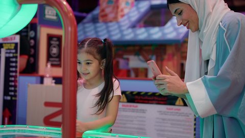 استخدام التقنيات الحديثه، امراه اماراتيه توثق اللحظات التذكاريه السعيده لابنتها ،طاولة مضيئة بألوان زاهية ، قضاء اوقات ممتعه مع العائله ، مراكز الالعاب الترفيهيه في الامارات، تصوير لطفلة عربية خليجية إماراتية ترتدي الزى الكاجوال ، تستمتع بلعب الهوكي الهوائي