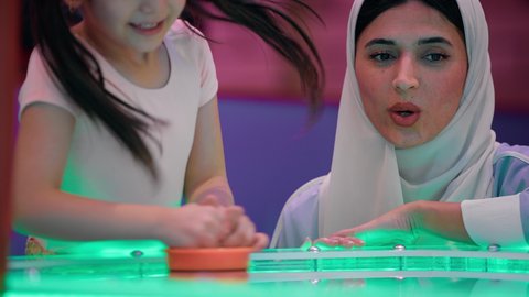 ايماءات المرح والحماس، قضاء اوقات ممتعه مع العائله ، مراكز الالعاب الترفيهيه في الامارات، تصوير لطفلة عربية خليجية إماراتية ترتدي الزى الكاجوال ، تستمتع بلعب الهوكي الهوائي برفقة والدتها ، طاولة مضيئة بألوان زاهية
