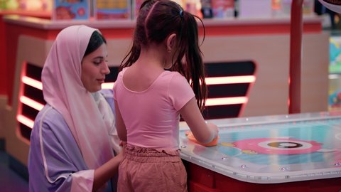 Spending fun time with the family, gestures of fun and enthusiasm, entertainment centers in the Emirates, a photo of an Emirati Gulf Arab girl wearing casual clothes, enjoying playing air hockey with her mother, a table illuminated with bright colors