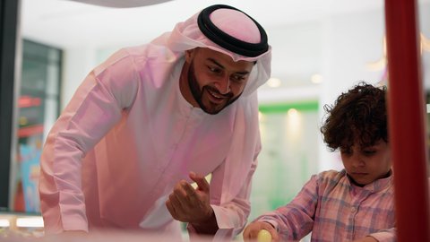 Entertainment centers in the Emirates, having a good time, an atmosphere full of fun and excitement, a photo of an Emirati Gulf Arab child wearing casual clothes, enjoying playing air hockey with his father, a table illuminated with bright colors