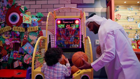 الأنشطة الرياضية الممتعة، أجواء مليئة بالمرح والحماس، تصوير لرجل عربي خليجي إماراتي يرتدي الكندورة والغترة يشارك أطفاله لحظات ممتعة في لعبة كرة السلة الإلكترونية فى أحد المراكز الترفيهية في الإمارات، منافسة مليئة بالتحدي والإثارة ، التفاعل بين أفراد العائلة.