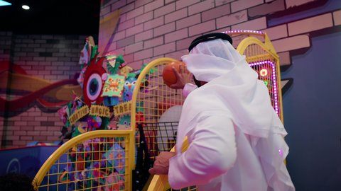 أجواء مليئة بالمرح والحماس، تصوير لرجل عربي خليجي إماراتي يرتدي الكندورة والغترة يشارك أطفاله لحظات ممتعة في لعبة كرة السلة الإلكترونية فى أحد المراكز الترفيهية في الإمارات، منافسة مليئة بالتحدي والإثارة ، الأنشطة الرياضية الممتعة، التفاعل بين أفراد العائلة.