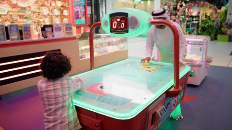 تصوير لرجل عربي خليجي اماراتي يرتدي الكندورة والغترة ، يلعب الهوكي الهوائي مع ابنه ، مراكز الالعاب الترفيهيه في الامارات ،الاستمتاع باللعب مع الابناء ، ايماءات التحدى والحماس ، طاولة مضيئة بألوان زاهية