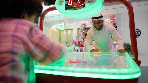 مراكز الالعاب الترفيهيه في الامارات ،الاستمتاع باللعب مع الابناء ، ايماءات التحدى والحماس ، تصوير لرجل عربي خليجي اماراتي يرتدي الكندورة والغترة ، يلعب الهوكي الهوائي مع ابنه ، طاولة مضيئة بألوان زاهية