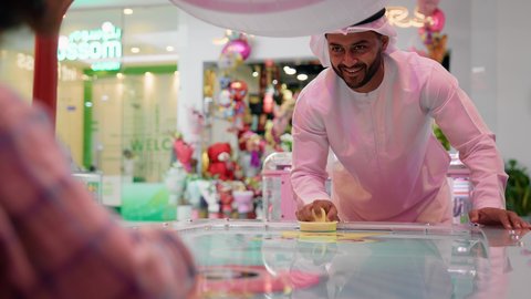 الاستمتاع باللعب مع الابناء ، ايماءات التحدى والحماس ، تصوير لرجل عربي خليجي اماراتي يرتدي الكندورة والغترة ، يلعب الهوكي الهوائي مع ابنه ، طاولة مضيئة بألوان زاهية ، مراكز الالعاب الترفيهيه في الامارات