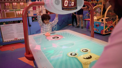 ايماءات الفوز والانتصار ، أجواء عائلية ممتعة ، تصوير لرجل عربي خليجي اماراتي يرتدي الكندورة والغترة ، يلعب الهوكي الهوائي مع ابنه ، طاولة مضيئة بألوان زاهية ، مراكز الالعاب الترفيهيه في الامارات