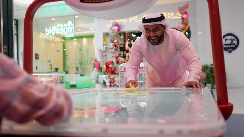 تعابير التحدي والحماس ، أجواء عائلية ممتعة ، تصوير لرجل عربي خليجي اماراتي يرتدي الكندورة والغترة ، يلعب الهوكي الهوائي مع ابنه ، طاولة مضيئة بألوان زاهية ، مراكز الالعاب الترفيهيه في الامارات