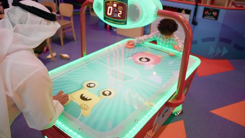 أجواء عائلية ممتعة ، تصوير لرجل عربي خليجي اماراتي يرتدي الكندورة والغترة ، يلعب الهوكي الهوائي مع ابنه ، طاولة مضيئة بألوان زاهية ، مراكز الالعاب الترفيهيه في الامارات  ، تعابير التحدي والحماس