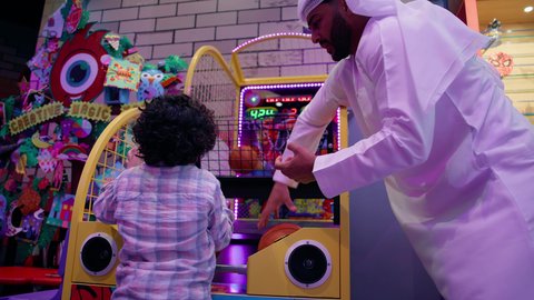 Entertainment games centers in the Emirates, combining modern technologies in children's games, spending fun times with the family, photography from the back to an Arab Gulf man, Ghamrat, wearing a ghutra and Kandoura shares his son game of basketball, spending fun time playing and focusing, enhancing mental skills