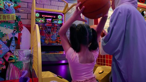 المتعة في مراكز الالعاب الترفيهيه في الامارات، دمج التقنيات الحديثه فى ألعاب الاطفال، تصوير من الخلف للسيدة عربية خليجية اماراتية محجبة ترتدي العباءة تقوم بمشاركة ابنتها لعبة رمي كرة السلة، قضاء وقت ممتع باللعب والتركيز، تعزيز المهارات الذهنية عند الابناء