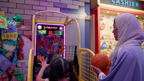 منافسة مليئة بالاثارة، أجواء حماسية مليئة بالمرح، تصوير من الخلف لفتاة سعودية تستمتع باللعب في مركز الالعاب الترفيهيه في الامارات، أم عربية خليجية اماراتية محجبة ترتدي العباءة تقوم بمشاركة ابنتها لعبة رمي كرة السلة، قضاء وقت ممتع باللعب والتركيز