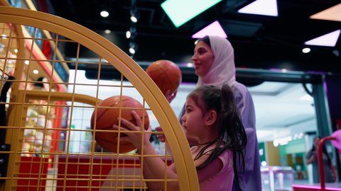 تحدي ومنافسات مليئة بالاثارة، أجواء حماسية مليئة بالمرح، فتاة سعودية تستمتع باللعب في مركز الالعاب الترفيهيه في الامارات، أم عربية خليجية اماراتية محجبة ترتدي العباءة تقوم بمشاركة ابنتها لعبة رمي كرة السلة، قضاء وقت ممتع باللعب والتركيز