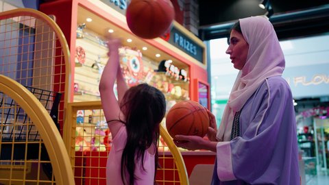 أجواء حماسية مليئة بالمرح، تعزيز المهارات الذهنية عند الابناء، فتاة سعودية تستمتع باللعب في مركز الالعاب الترفيهيه في الامارات، أم عربية خليجية اماراتية محجبة ترتدي العباءة تقوم بمشاركة ابنتها لعبة رمي كرة السلة، قضاء وقت ممتع باللعب والتركيز