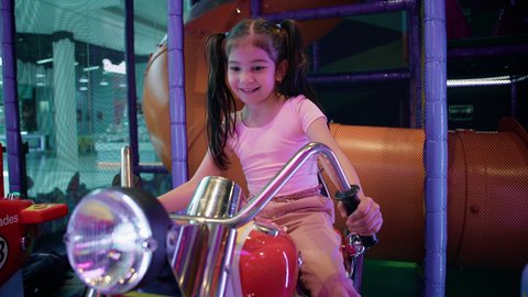 النظر إلى الكاميرا بايماءات السعادة والمتعة، العاب الاركيد الالكترونيه المتطورة، الاستمتاع بقضاء الاجازة الصيفيه في احدى الصالات في الأماكن الترفيهية في الامارات، فتاة عربية خليجية اماراتية ترتدي الزي الكاجوال وتقوم بركوب الدباب الكهربائي