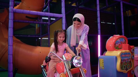 أم عربية خليجية سعودية ترتدي عباءة تقوم بمشاركة طفلتها لحظات المرح، المتعة في احدى الصالات الترفيهية في الامارات، ايماءات السعادة والسرور، فتاة عربية خليجية اماراتية ترتدي الزي الكاجوال وتقوم بركوب الدباب الكهربائي