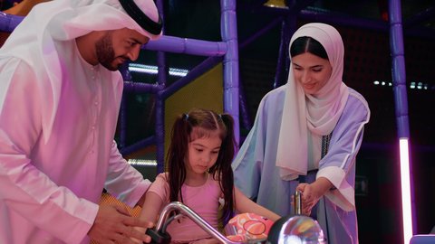 قضاء اوقات عائلية ممتعة، مشاركة الأب والأم طفلتهما لحظات المرح، الاستمتاع بقضاء الاجازة الصيفيه في احدى الصالات في الأماكن الترفيهية في الامارات، ايماءات السعادة والسرور، فتاة عربية خليجية اماراتية ترتدي الزي الكاجوال وتقوم بركوب الدباب الكهربائي